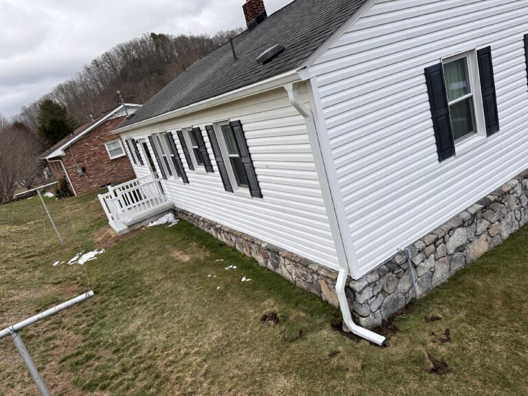 Flag Gutter Services Installing Gutters On A White House With Black Shutters And A Stone Foundation On A Grassy Area